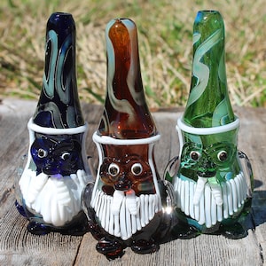 May include: Three glass pipes shaped like gnomes with white beards and hats. The pipes are decorated with swirls of blue, green, and brown glass.