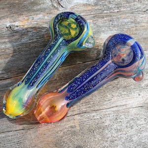 May include: Two glass pipes with colorful designs. One pipe has a blue and green body with yellow accents, while the other is blue and orange. Both pipes have a bowl and a stem, and are set against a wooden background.