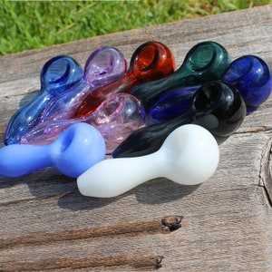 May include: A set of nine glass smoking pipes in various colors, including blue, pink, red, green, and white. The pipes are arranged in a semi-circle on a wooden surface.