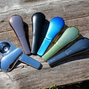 Puede incluir: Un conjunto de siete pipas para fumar de metal en varios colores, incluyendo negro, azul, verde, dorado y plateado. Las pipas están dispuestas en forma de abanico sobre una superficie de madera.