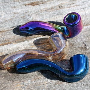 May include: Three glass smoking pipes in different colors and shapes. The pipes are iridescent, with a blue, purple, and brown finish. The pipes are arranged on a wooden surface.