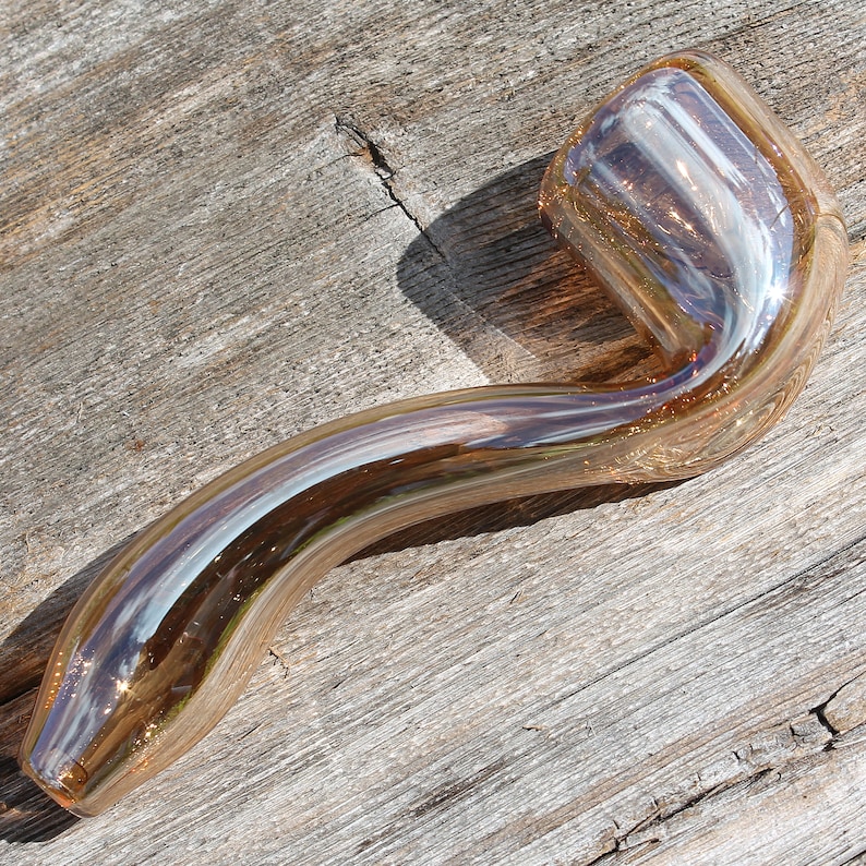 May include: A glass pipe with a curved stem and a rounded bowl. The pipe is made of clear glass with a light brown tint and has a iridescent finish.