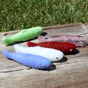 Puede incluir: Cinco figuras de peces de vidrio de colores verde, morado, rojo, blanco y azul, cada uno con un pequeño ojo negro. Los peces están dispuestos sobre una superficie de madera envejecida, con un fondo de hierba.