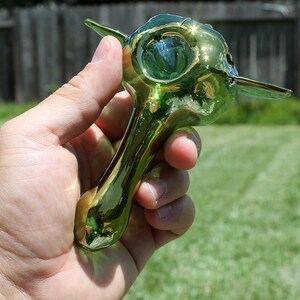 K&ouml;nnte beinhalten: Eine gr&uuml;ne Glaspfeife mit einem einzigartigen Design. Die Pfeife hat einen breiten Kopf und einen langen, d&uuml;nnen Schaft. Das Glas ist schillernd und hat einen goldenen Schimmer.