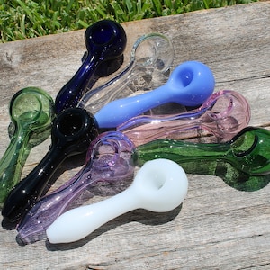 May include: A collection of nine glass smoking pipes in various colors, including green, blue, purple, and white. The pipes are arranged in a fan shape on a wooden surface.