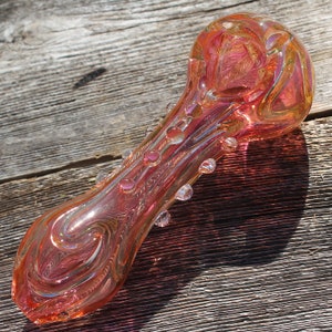 May include: A pink glass pipe with a swirl design and clear glass accents. The pipe is made of thick glass and has a rounded bowl.