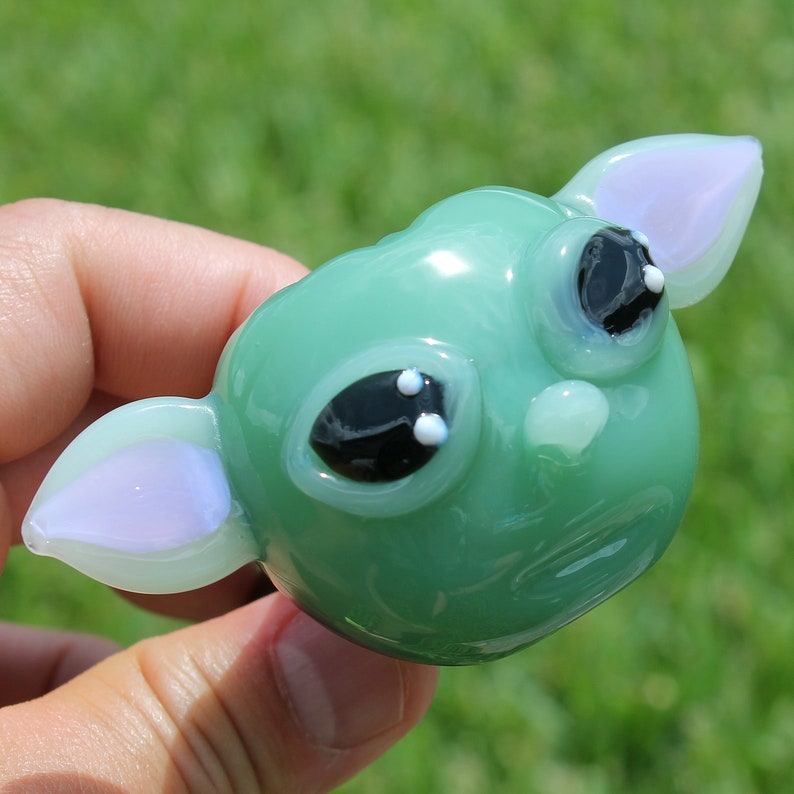 K&ouml;nnte beinhalten: Eine gr&uuml;ne Glasfigur eines Charakters mit gro&szlig;en Augen und zwei kleinen Ohren. Die Figur ist in einem hellgr&uuml;nen und wei&szlig;en Farbschema gehalten.