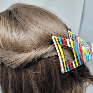 May include: A colorful hair claw clip with a geometric design. The clip features a clear base with vertical stripes in yellow, red, blue, and black. The clip is holding back brown hair, showcasing the accessory's design and function.
