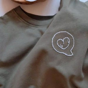 May include: A green t-shirt with a white embroidered heart in a speech bubble.