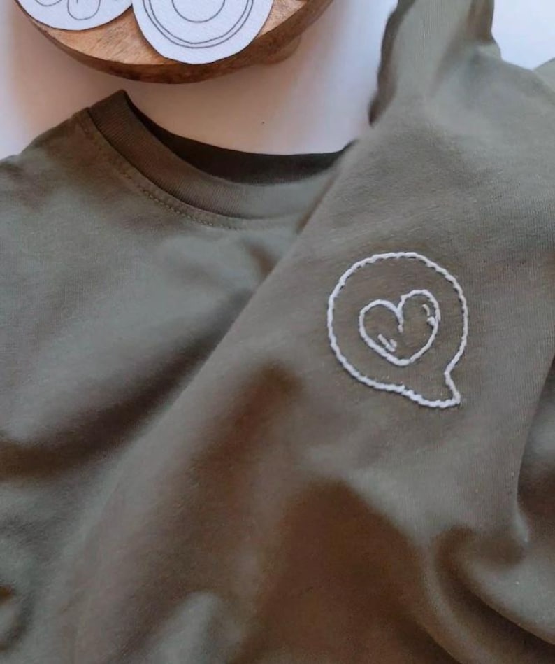 May include: A green t-shirt with a white embroidered heart in a speech bubble.