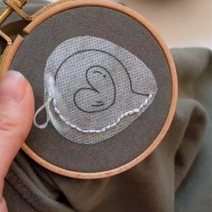 May include: A close-up of a green fabric with a white heart-shaped design embroidered on it. The fabric is stretched over a wooden embroidery hoop.
