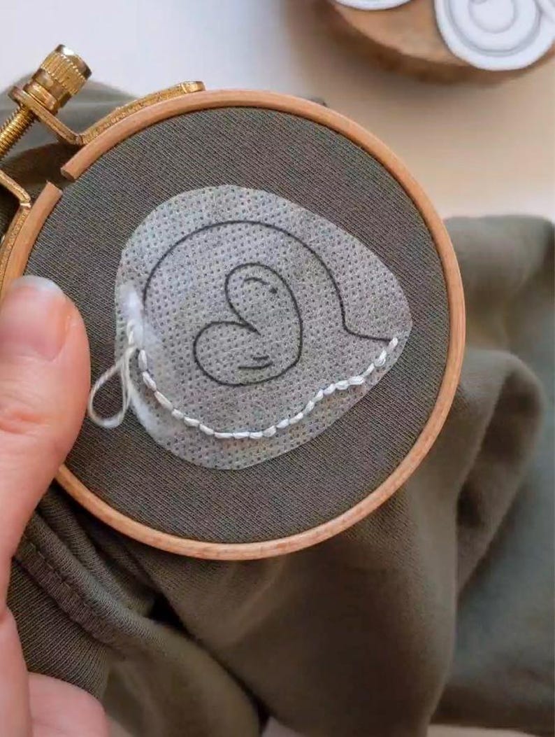 May include: A close-up of a green fabric with a white heart-shaped design embroidered on it. The fabric is stretched over a wooden embroidery hoop.
