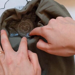 May include: A close-up of a hand pressing a heart-shaped iron-on patch onto a green t-shirt. The patch is surrounded by a clear adhesive.