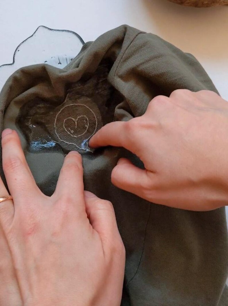 May include: A close-up of a hand pressing a heart-shaped iron-on patch onto a green t-shirt. The patch is surrounded by a clear adhesive.