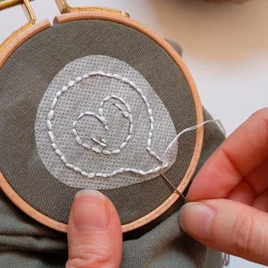 May include: A close-up of a person embroidering a white heart shape on a green fabric using a wooden embroidery hoop.