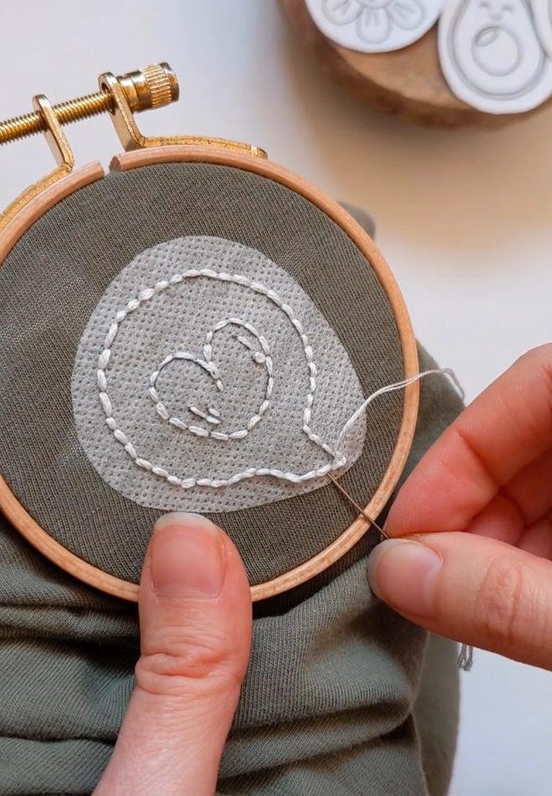 May include: A close-up of a person embroidering a white heart shape on a green fabric using a wooden embroidery hoop.
