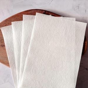 May include: Four white, rectangular, textured paper sheets stacked on top of each other on a wooden surface.