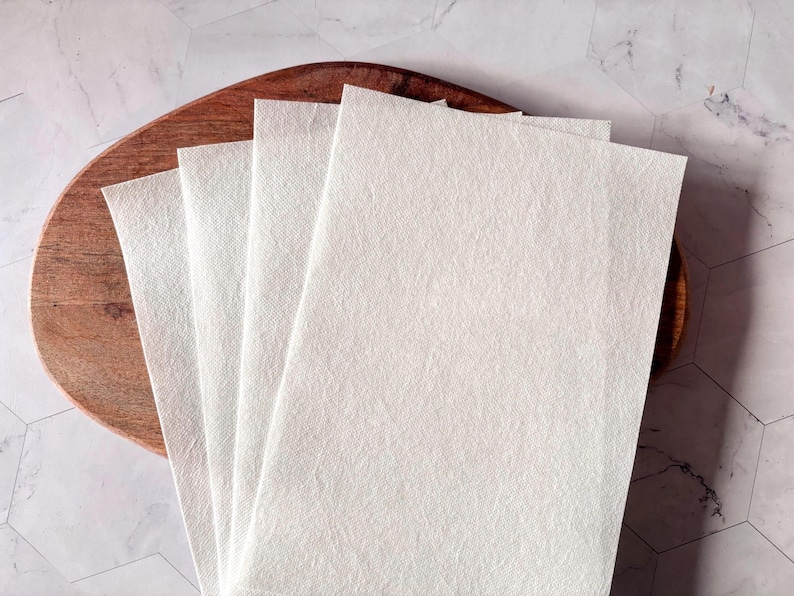 May include: Four white, rectangular, textured paper sheets stacked on top of each other on a wooden surface.