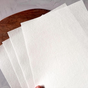 May include: A stack of white paper sheets with a textured surface. The sheets are folded in half and are lying on a wooden surface.