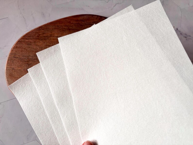 May include: A stack of white paper sheets with a textured surface. The sheets are folded in half and are lying on a wooden surface.