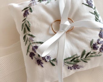 May include: White ring pillow with a lavender embroidery design and a white satin ribbon tied around it. Two gold wedding bands are placed on the pillow.