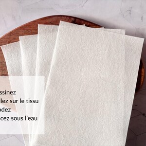 May include: Four white fabric squares stacked on a wooden surface. The text "Dessinez Collez sur le tissu Brodez Rincez sous l'eau" is printed on the bottom square.