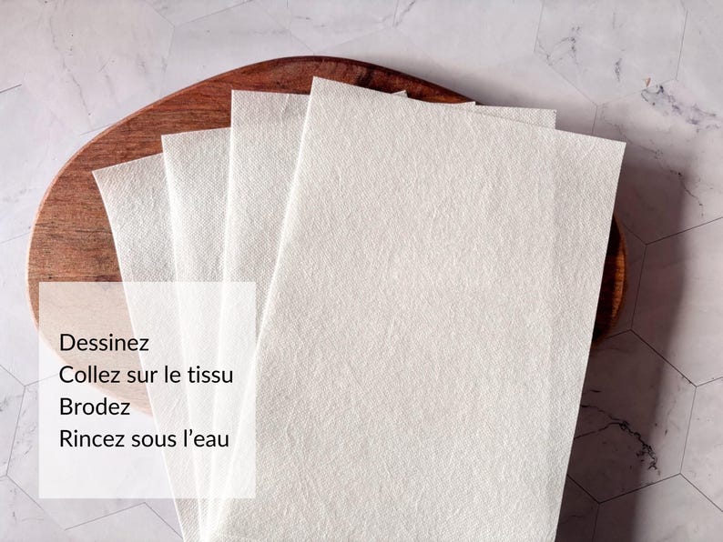 May include: Four white fabric squares stacked on a wooden surface. The text "Dessinez Collez sur le tissu Brodez Rincez sous l'eau" is printed on the bottom square.