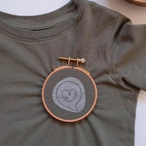 May include: A green t-shirt with a small embroidery hoop featuring a white speech bubble with a heart inside.