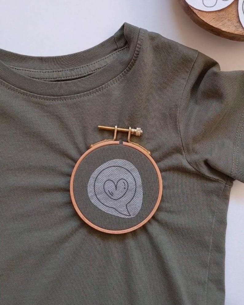 May include: A green t-shirt with a small embroidery hoop featuring a white speech bubble with a heart inside.