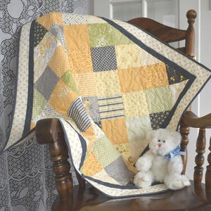 May include: A yellow, green, and gray patchwork quilt with a white border and a gray trim. The quilt is folded and draped over a wooden rocking chair with a white teddy bear sitting on top of it.