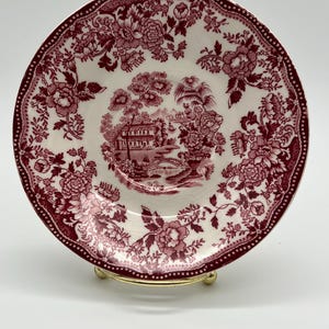 May include: A round ceramic saucer featuring a deep red transferware pattern on a white background. The center depicts a landscape with a building and bridge, while the border is decorated with intricate floral motifs and a dotted edge design.