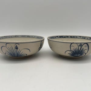 以下が含まれることがあります： 青い花柄のオフホワイトのセラミックボウルが2つ。各ボウルには青い装飾的なボーダーと、側面に様式化された青い花のモチーフがあります。ボウルは、おそらく食べ物を提供するものです。