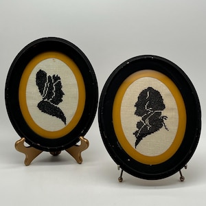 May include: Two framed silhouette portraits. Each oval frame is black with a gold inner border. The portraits are black and white, depicting profiles of people. The frames are displayed on decorative stands.