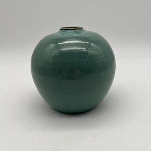 Puede incluir: Un jarrón de cerámica redondo, de color verde azulado, con una abertura estrecha y un borde marrón oscuro. El jarrón tiene un acabado brillante y una forma lisa y redondeada. El fondo es blanco liso.