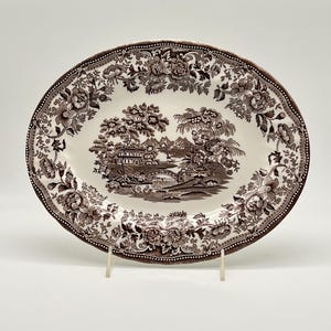 May include: An oval ceramic platter featuring a dark brown transferware pattern on a cream background. The center depicts a landscape with a bridge, trees, and buildings, while the wide rim is decorated with a dense floral and leaf motif.