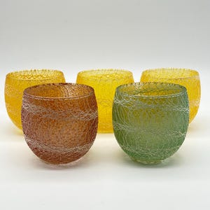 May include: Five colorful, round glass bowls. The bowls are in shades of yellow, brown, and green, with a textured, web-like design. The bowls are arranged on a white background.