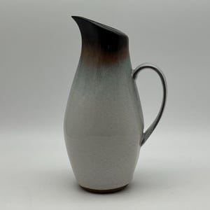 May include: A white ceramic pitcher with a brown and black glaze. The pitcher has a rounded body and a curved handle.