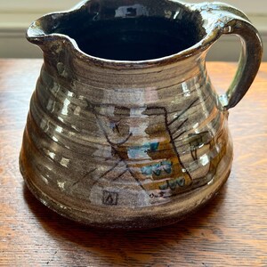 May include: A brown ceramic pitcher with a stylized animal design in brown, blue, and yellow. The pitcher has a wide mouth and a handle.