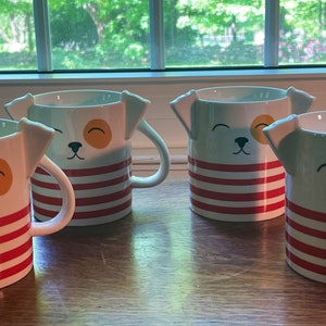 Puede incluir: Cuatro tazas de cerámica blanca con rayas rojas y blancas y una cara de perro de dibujos animados con ojos amarillos. Las tazas tienen pequeñas orejas que sobresalen de los lados.