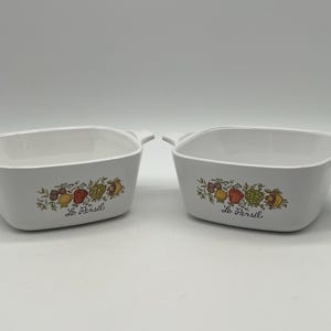 May include: Two white rectangular baking dishes with handles. Each dish features a colorful floral and vegetable design with the text "Le Persil". The design includes red, yellow, and green elements. The dishes are empty and sit on a white surface.