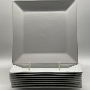 Op de afbeelding: Een stapel witte vierkante borden. De borden hebben een eenvoudig, modern ontwerp met licht afgeronde hoeken. De borden zijn netjes opgestapeld, met één bord bovenop en verschillende andere eronder.