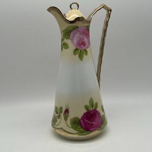 May include: A vintage porcelain pitcher with a cream-colored body, adorned with pink roses and green leaves. The pitcher features a gold-trimmed handle and spout, and a matching lid with a gold ring. The design is a classic floral pattern.