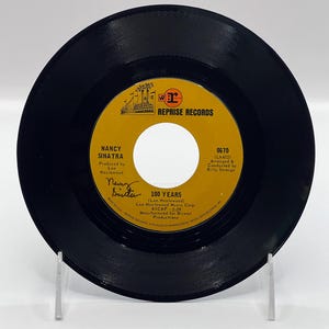 Peut inclure: Un disque vinyle noir avec une étiquette dorée. L'étiquette présente le texte "Reprise Records", "Nancy Sinatra", "100 Years", "Lee Hazlewood", "ASCAP 2:29", "0670 (L6402)", "Arrangé et dirigé par Billy Strange", et "Fabriqué pour Bristol Productions".