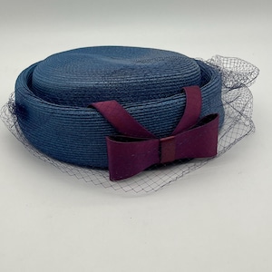 Puede incluir: Un sombrero azul marino con un diseño tejido estructurado. El sombrero presenta un lazo burdeos oscuro y un velo delicado y transparente. Un accesorio de estilo vintage.