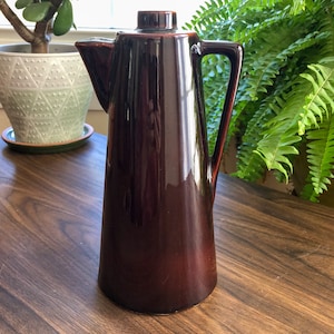 Puede incluir: Una cafetera de cerámica marrón oscuro con una forma alta y cónica y un asa en el lado derecho. La cafetera tiene un acabado liso y brillante.