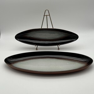 Może przedstawiać: Dwa owalne ceramiczne talerze do serwowania. Górny talerz jest czarny z brązowymi i białymi smugami. Dolny talerz jest biały z brązową obwódką i czarnymi smugami.