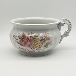 May include: A white ceramic chamber pot featuring a decorative floral pattern with pink and yellow roses on the front. The vessel has a wide, rounded bowl shape, a flared rim, and a single ornate, curved handle attached to the right side.