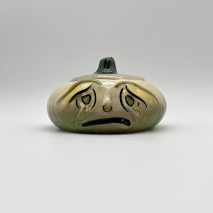 May include: A ceramic lidded jar shaped like a sad, crying onion with a pale green and cream-colored body. The onion features hand-painted black outlines for the eyes, eyebrows, and mouth, with green tear drops and a dark green stem on the lid.