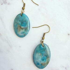 Op de afbeelding: Een paar hangende oorbellen met ovaalvormige hangers met een gemarmerd patroon in tinten turquoise, blauw en wit. De hangers hangen aan antiek messingkleurige metalen vishaakjes met een klein spiraal- en kraaldetail.