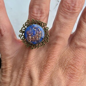 Op de afbeelding: Een ring met een ronde blauwe hars cabochon met ingebedde koperkleurige metaalvlokken. De hars is gezet in een antiek bronskleurige metalen filigraan zetting met een ingewikkeld, wervelend opengewerkt ontwerp rondom het midden.